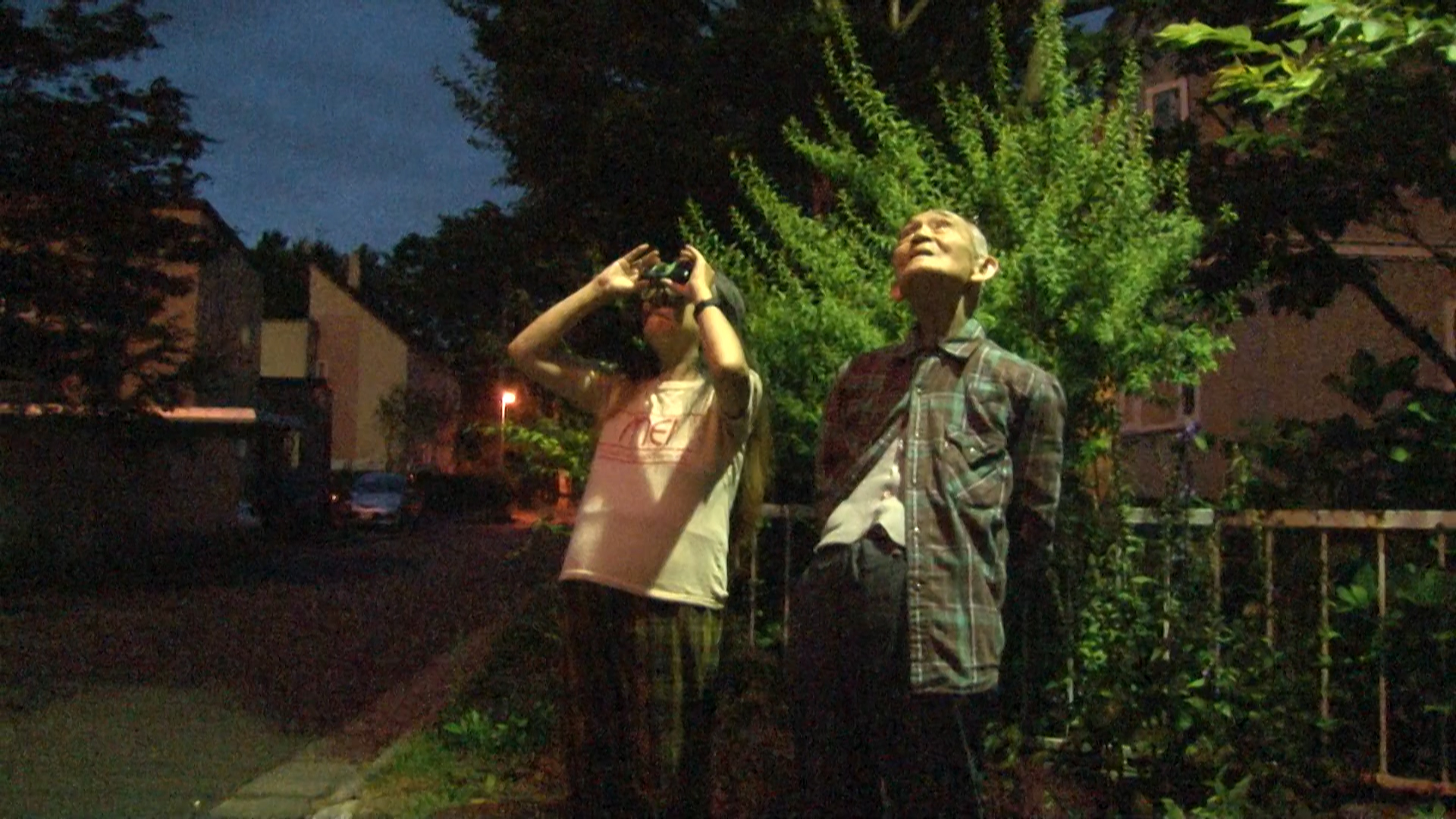 映画の場面写真。夕暮れの屋外で双眼鏡を手にする人